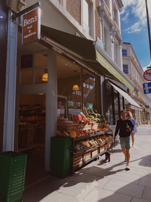 Exterior view of a small grocery store with a sign reading 'Bens fruit & vegetables,' located on a busy urban street. The store has large glass windows revealing the interior, which is illuminated by hanging pendant lights. Outside, on the sidewalk, there is a wooden display shelf filled with fresh produce such as watermelons, melons, and other fruits, arranged in baskets and trays. A green plastic crate is positioned at the corner of the storefront, and the store’s entrance is partially visible to the left. The surrounding environment includes multi-story buildings with neutral-colored facades and other storefronts, some with awnings. The scene shows pedestrians walking by, including a man in a black shirt and shorts carrying a backpack, walking past the store in daylight, with a partly cloudy sky overhead. This setting portrays a typical shopfront in a busy city area, relevant for discussions regarding local retail relocations and urban moving services by Kennington Movers.