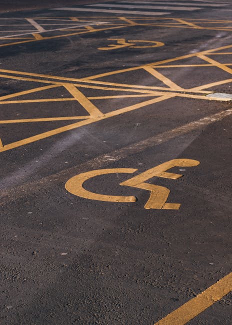 A close-up view of an asphalt parking lot surface displaying yellow painted markings for accessible parking spaces and designated parking bays. The prominent accessible parking symbol, featuring a wheelchair icon, is painted on the ground in the foreground, with additional yellow lines marking individual parking spots and yellow diagonal hatchings visible further back. The surface shows slight wear and tire marks, indicating recent or frequent vehicle use. This scene illustrates a typical outdoor parking area, relevant to house removals and logistics work, such as loading and unloading furniture or packing materials. The image emphasizes the importance of understanding parking regulations, including permit rules by Lambeth Council, for efficient home relocation operations. Kennington Movers occasionally operate in such environments, ensuring compliance with local parking requirements during packing, loading, and furniture transport processes.
