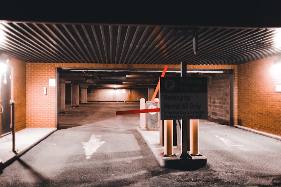 Interior view of a multi-storey underground parking garage with brick walls and a black, ribbed ceiling. A closed gate with orange and red barrier arms blocks vehicle access, supported by concrete and metal pillars. A parking sign indicating parking lot 60 and permit restrictions is mounted on the central pillar, with partially visible text reading 'Parking by Permit 60 Only.' The pavement beneath the barriers shows a white arrow painted on the ground pointing forward, guiding vehicles into the parking area. The scene is lit by overhead lighting, creating an industrial atmosphere suitable for vehicle and home relocation logistics, and reflects the type of secure parking environment often used during house removals, facilitated by local authorities such as Lambeth Council. The space appears clean and organized, prepared for loading or unloading furniture and packing materials associated with removals or moving services, including visible protective barriers for safe traffic control.