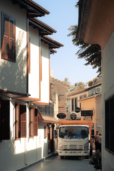 A narrow street in a residential area with white buildings featuring wooden shutters and overhanging roofs, where a flatbed trolley loaded with cardboard boxes and a large wrapped piece of furniture is positioned near the entrance of a building. A Kennington Movers van is parked further along the street, with a person visible nearby, likely involved in the loading process of items for a home relocation. The scene is set during daylight with natural light illuminating the area, and trees partially visible above the buildings. The environment suggests an active furniture transport and packing operation as part of professional removals services focused on house moves and transport logistics.