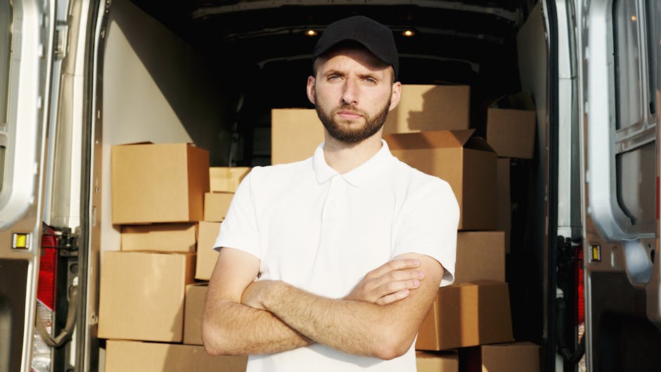 A young man with a beard, wearing a black cap and a white polo shirt, stands inside the open rear door of a large moving van filled with stacked cardboard boxes. The boxes vary in size and are secured