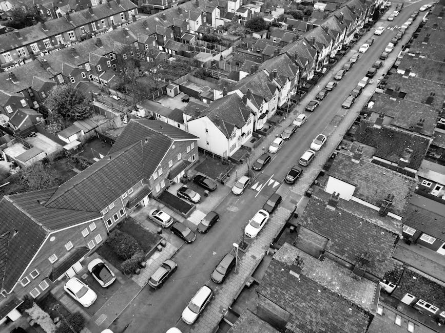 An aerial black-and-white photograph displays a residential street with closely packed terraced houses and semi-detached homes, typical of an urban neighbourhood. The street is lined with parked cars on both sides, predominantly compact and medium-sized vehicles, with some occupying driveways of individual houses. The road features a clear central divider with striped markings and directional arrows. Sidewalks run alongside the houses, where pedestrians could walk and residents may access their properties. In the foreground, a house with a pitched roof and small front garden is visible, and nearby, a driveway with additional parked cars leads to a garage or storage area. A section of the street appears to be in the process of a home relocation or furniture transport operation, with materials such as cardboard boxes, packing wrap, and possibly furniture being loaded onto a nearby van. A Kennington Movers vehicle is parked on the side of the road, facilitating an efficient loading process in this urban setting, suitable for house removals within narrow streets like those described in the Kennington Park house removals guide for narrow streets.