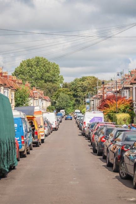 A residential street scene showing a narrow pavement lined with parked cars on both sides, including sedans, hatchbacks, and vans, in front of terraced houses with brick facades, small front gardens, and chimneys. The street is relatively quiet, with a few vehicles visible in the distance. Overhead, power lines cross the cloudy sky. In the foreground, a portion of a large moving van from Kennington Movers is visible, with the side door open, and packing blankets and cardboard boxes arranged nearby, indicating a home relocation process. The scene captures the typical urban environment for house removals, highlighting the challenges of narrow streets and close parking when transporting furniture and belongings. Some of the vehicles are covered with protective tarps or plastic wraps to prevent damage during loading or transport, emphasizing careful planning for furniture transport and moving logistics in densely built residential areas. The overall scene reflects a professional moving company's preparation for house removals on a typical street, blending elements of packing, loading, and vehicle loading procedures.