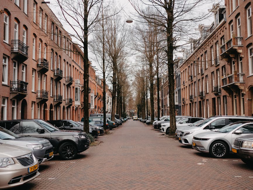 An aerial black-and-white photograph displays a residential street with closely packed terraced houses and semi-detached homes, typical of an urban neighbourhood. The street is lined with parked cars on both sides, predominantly compact and medium-sized vehicles, with some occupying driveways of individual houses. The road features a clear central divider with striped markings and directional arrows. Sidewalks run alongside the houses, where pedestrians could walk and residents may access their properties. In the foreground, a house with a pitched roof and small front garden is visible, and nearby, a driveway with additional parked cars leads to a garage or storage area. A section of the street appears to be in the process of a home relocation or furniture transport operation, with materials such as cardboard boxes, packing wrap, and possibly furniture being loaded onto a nearby van. A Kennington Movers vehicle is parked on the side of the road, facilitating an efficient loading process in this urban setting, suitable for house removals within narrow streets like those described in the Kennington Park house removals guide for narrow streets.