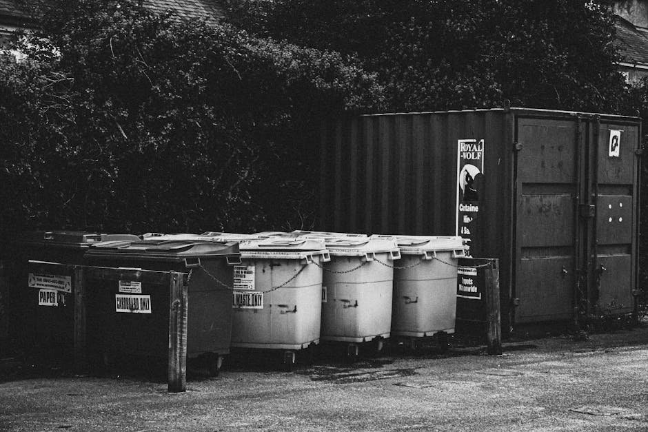 A row of waste disposal bins and containers positioned outside a residential property, with some labeled for specific refuse types such as paper, general waste, and household waste, situated on a paved area next to a black metal barrier and a large dark shipping container. The bins are made of plastic, with lids closed, and are arranged in a line parallel to the building, which is partially visible in the background, shaded by overhanging trees and foliage. The scene depicts the process of waste collection related to home relocation or moving services, with the bins prepared for rubbish disposal in accordance with local regulations, as handled by Kennington Movers in compliance with Lambeth Council's rules for waste and move-related disposal.