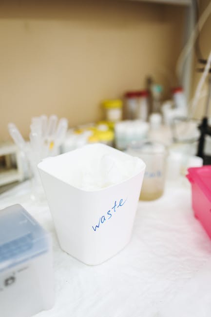 A white plastic container labeled 'waste' in blue handwriting sits on a white surface, surrounded by various small bottles, jars, and containers, many with yellow caps, indicating laboratory or cleaning supplies. In the background, shelves hold additional bottles, containers, and packaging materials commonly used in home or office environments. The scene appears to be set indoors, possibly in a packing or sorting area used during a home relocation or moving process managed by Kennington Movers. The clear organization and labeling support efficient waste disposal, which is part of the moving and packing process involved in furniture transport and house removals, particularly when adhering to local waste disposal rules in Lambeth.