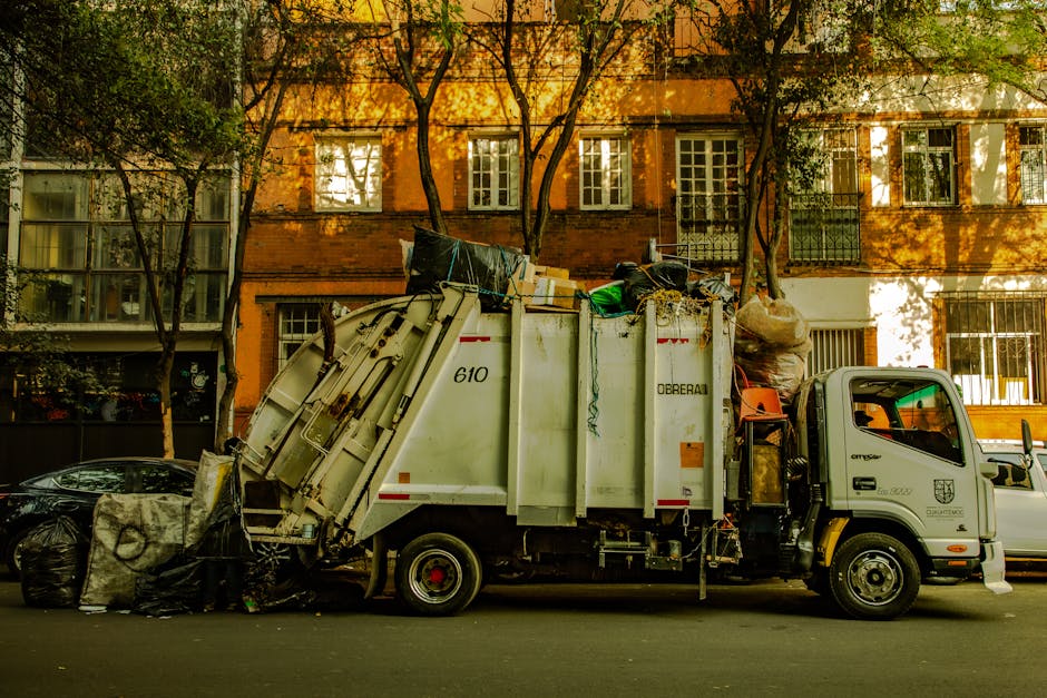 A row of waste disposal bins and containers positioned outside a residential property, with some labeled for specific refuse types such as paper, general waste, and household waste, situated on a paved area next to a black metal barrier and a large dark shipping container. The bins are made of plastic, with lids closed, and are arranged in a line parallel to the building, which is partially visible in the background, shaded by overhanging trees and foliage. The scene depicts the process of waste collection related to home relocation or moving services, with the bins prepared for rubbish disposal in accordance with local regulations, as handled by Kennington Movers in compliance with Lambeth Council's rules for waste and move-related disposal.