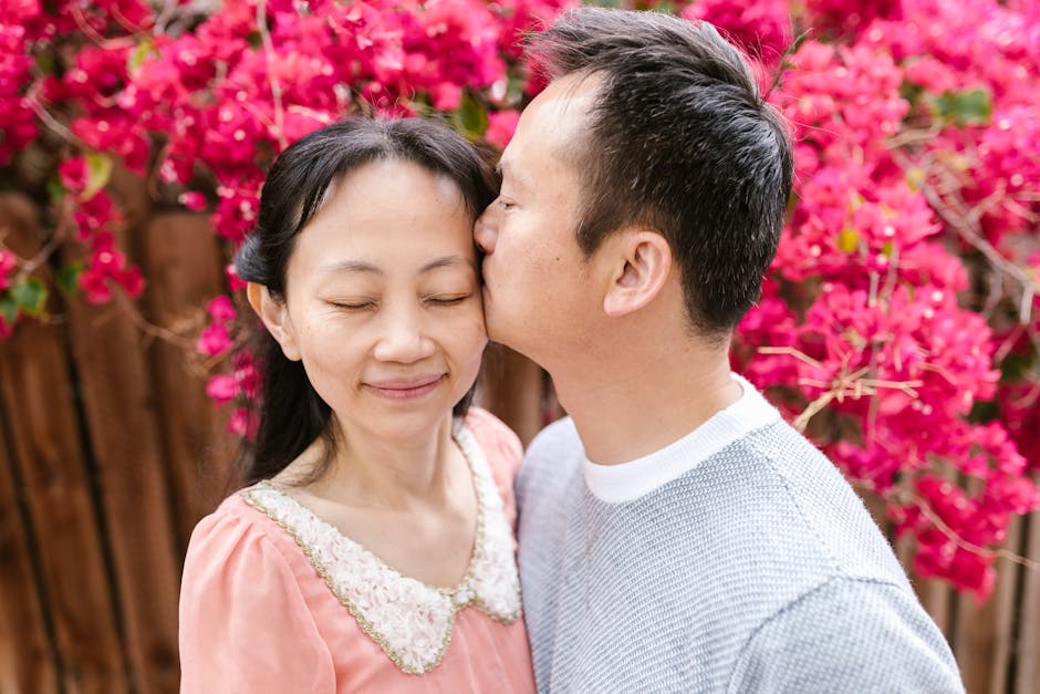 A man giving a gentle kiss on the forehead to a woman with closed eyes, both standing outdoors in front of a vibrant pink flowering shrub, with a wooden fence visible behind them. The woman is wearing