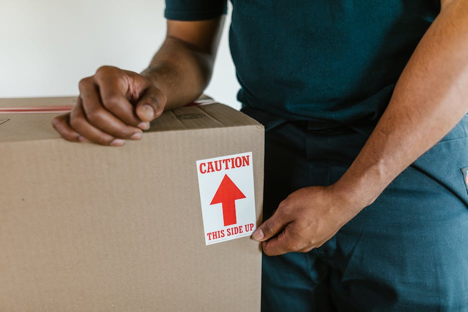 Close-up of a person wearing a dark blue shirt and dark pants, holding a large cardboard box with both hands. The box is sealed and has a red and white caution sticker prominently displayed on the side, indicating 'CAUTION THIS SIDE UP' with an upward-pointing arrow. The individual appears to be preparing the box for a home relocation or furniture transport, with the background showing an indoor environment. The image captures the careful handling typical during packing and loading processes involved in professional removals. Occasionally, Kennington Movers' services include packing, moving, and loading systems as part of comprehensive house removals in SE11 and surrounding areas, emphasizing safe handling and secure transport of household items.
